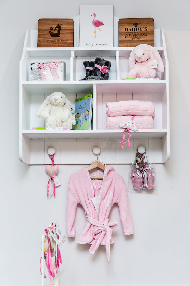 White Lyra wall shelf with open cubbies, pigeon holes, tongue-and-groove detail and three rounded hooks, styled for nursery or kids’ room
