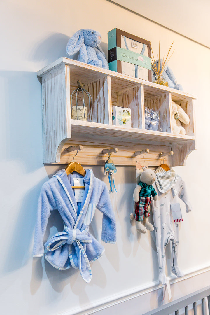 Distressed white Orchid wall shelf with four cubbies and three hooks, styled with baby clothes and toys, ideal for nursery storage
