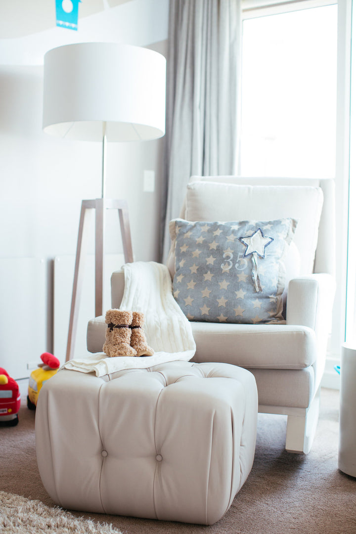 White Jayden rocking chair styled with a star-print cushion, soft knit throw, and paired with a tufted ottoman featuring a teddy bear and baby booties, set in a cozy nursery corner.