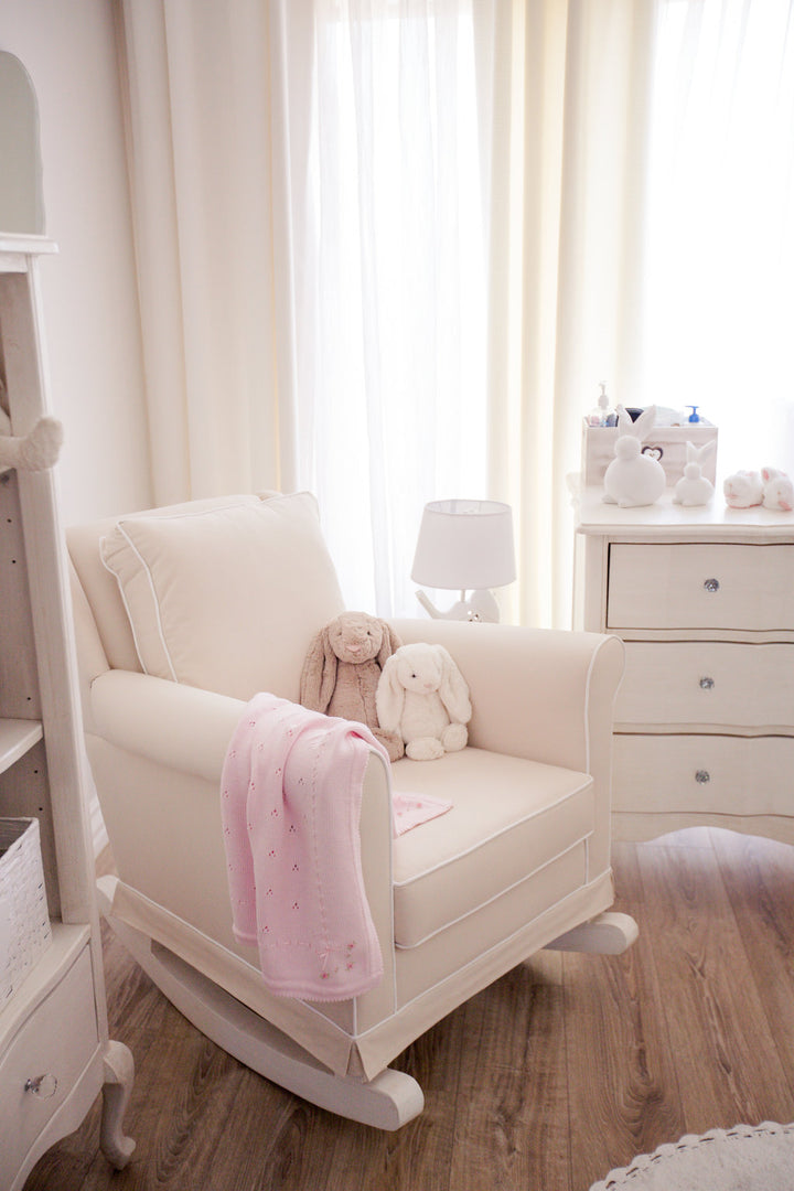 Tres Chic Rocking Chair in soft ivory fabric with natural wooden rocking legs, styled in a serene nursery with plush bunny toys and a pink baby blanket draped over the arm.