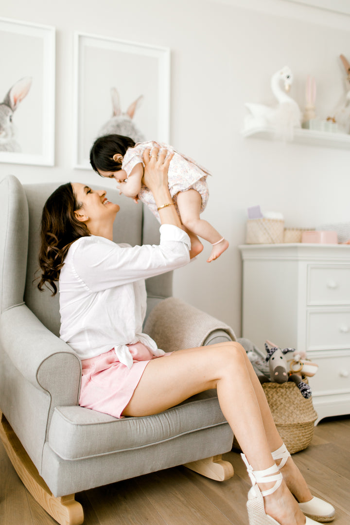 A mother sits with her baby on the Valentina Rocking Chair in a soft grey fabric with natural oak rocking legs, styled in a serene nursery with white furniture, animal artwork, and a small dog in the foreground.